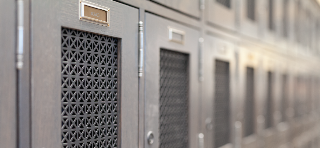 Image of a row of multiple wine lockers. They are a light grey wood grain with a grate in the middle of each door. They have customized gold plaques above the grates with the name of the individual or group who owns the wine locker.